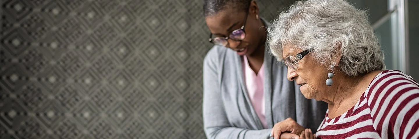 Eine Pflegerin hilft einer älteren Frau, die Treppe hinab zu steigen. Im Hintergrund sind ein Treppengeländer und eine gemusterte Tapete zu sehen.