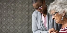 Eine Pflegerin hilft einer älteren Frau, die Treppe hinab zu steigen. Im Hintergrund sind ein Treppengeländer und eine gemusterte Tapete zu sehen.