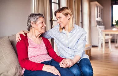 Eine alte Frau sitzt in einem Wohnzimmer auf der Couch. Neben ihr sitzt eine junge Frau. Beide schauen sich an, halten sich gegenseitig die Hand und lächeln einander an.