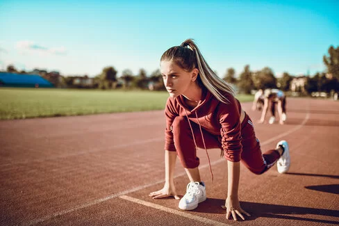 Eine junge Frau steht im tiefen Ausfallschritt auf einem Sportplatz, bereit für den Laufstart. Die AOK NordWest veranstaltet jährlich den Laufabzeichen-Wettbwerb "Laufwunder".