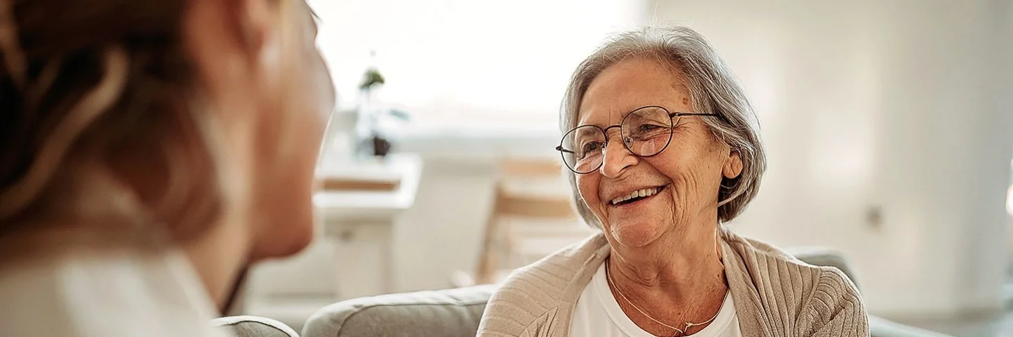 Ältere Frau sitzt lächelnd auf einem Sofa und spricht mit einer jüngeren Person, die ihr gegenübersitzt. Die Szene wirkt warm und vertraut.