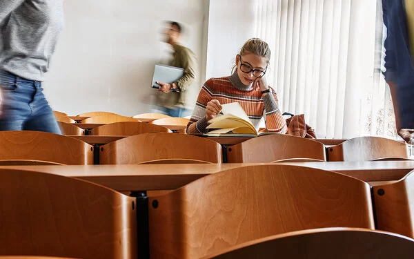 Studentin sitzt in einer Stuhlreihe eines Hörsaals und blättert durch ein Buch auf dem Pult.