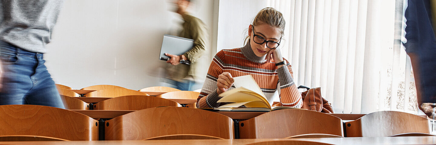 Studentin sitzt in einer Stuhlreihe eines Hörsaals und blättert durch ein Buch auf dem Pult.