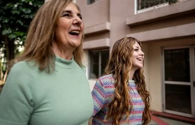 Nahaufnahme zweier Frauen, die nebeneinander gehen und lachen; städtischer Innenhof im Hintergrund.