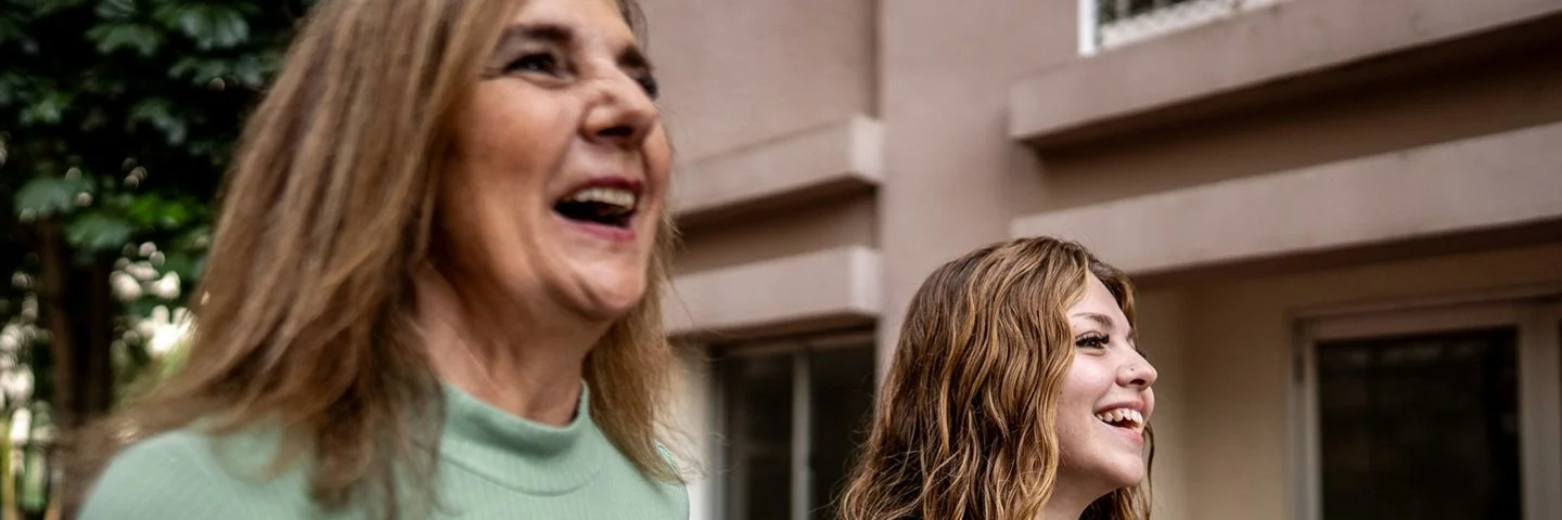 Nahaufnahme zweier Frauen, die nebeneinander gehen und lachen; städtischer Innenhof im Hintergrund.