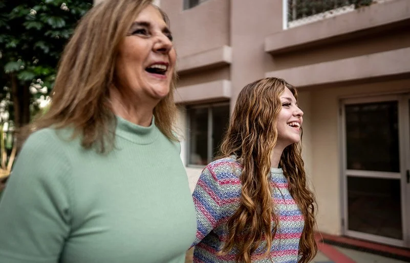 Nahaufnahme zweier Frauen, die nebeneinander gehen und lachen; städtischer Innenhof im Hintergrund.