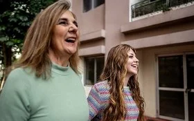 Nahaufnahme zweier Frauen, die nebeneinander gehen und lachen; städtischer Innenhof im Hintergrund.