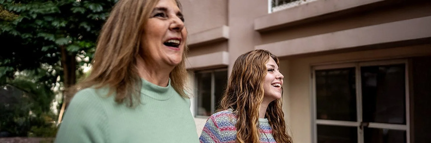 Nahaufnahme zweier Frauen, die nebeneinander gehen und lachen; städtischer Innenhof im Hintergrund.