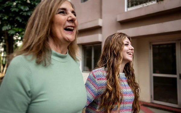 Nahaufnahme zweier Frauen, die nebeneinander gehen und lachen; städtischer Innenhof im Hintergrund.