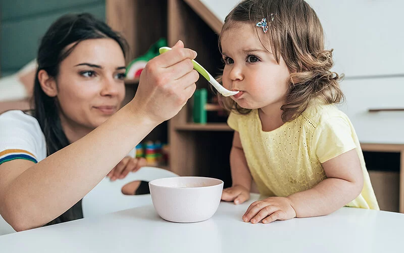 Ein kleines Mädchen sitzt am Tisch und wird von der Mutter mit einem Löffel gefüttert.