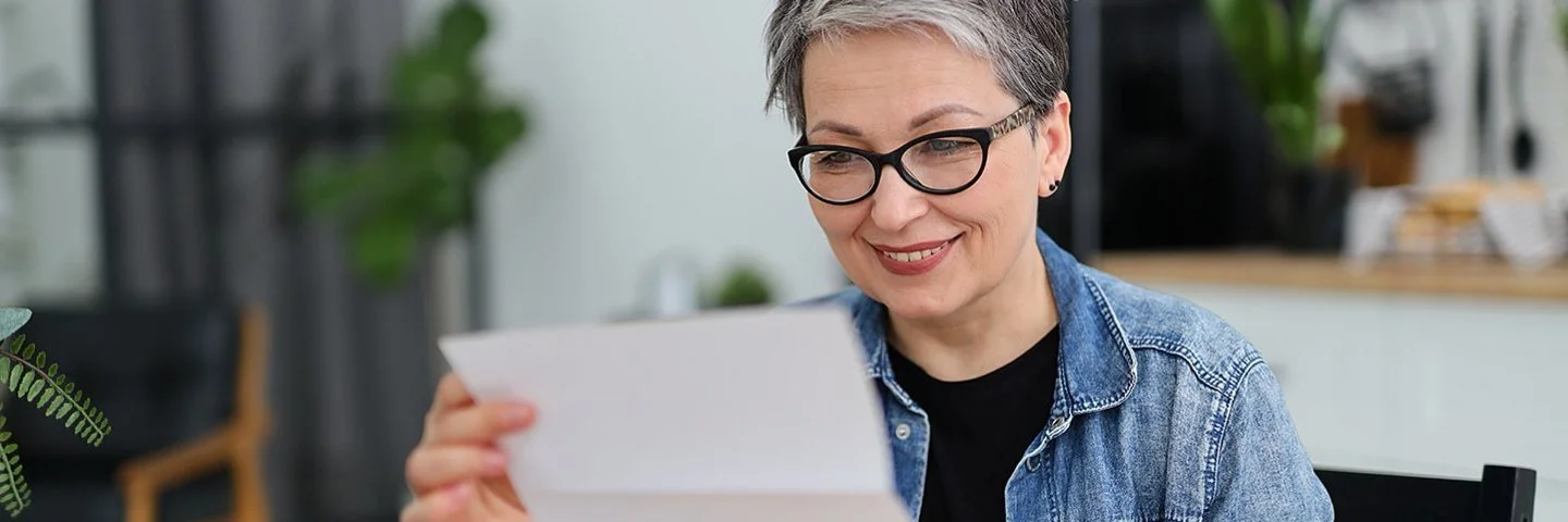 Eine Frau mit kurzen Haaren sitzt in einer Küche, ein geöffneter Briefumschlag liegt auf dem Tisch. Sie blickt auf einen Brief, den sie in ihren Händen hält.