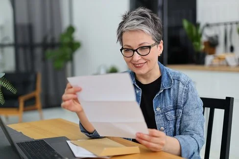 Eine Frau mit kurzen Haaren sitzt in einer Küche, ein geöffneter Briefumschlag liegt auf dem Tisch. Sie blickt auf einen Brief, den sie in ihren Händen hält.