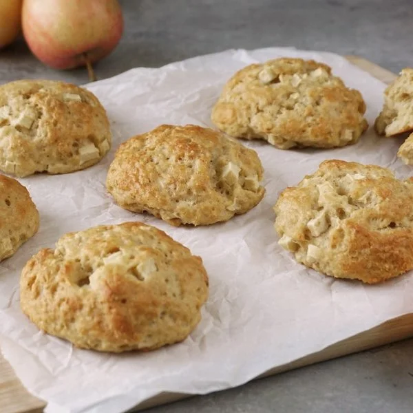 Apfel-Quark-Haferflocken-Brötchen auf einem Holzbrett mit Pergamentpapier. Im Vordergrund auf grauem Untergrund ein halbierter Apfel, im Hintergrund Äpfel und Zimtstangen.