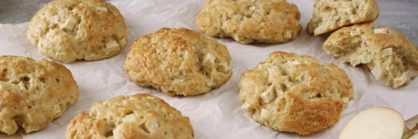 Apfel-Quark-Haferflocken-Brötchen auf einem Holzbrett mit Pergamentpapier. Im Vordergrund auf grauem Untergrund ein halbierter Apfel, im Hintergrund Äpfel und Zimtstangen.