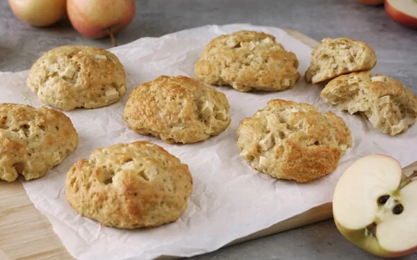 Apfel-Quark-Haferflocken-Brötchen auf einem Holzbrett mit Pergamentpapier. Im Vordergrund auf grauem Untergrund ein halbierter Apfel, im Hintergrund Äpfel und Zimtstangen.