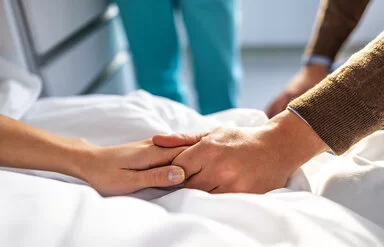 Eine Frau liegt in einem Krankenbett, ein Mann sitzt neben ihr. Zu sehen sind jedoch nur die Hände der beiden, die sich festhalten. Im Hintergrund eine medizinische Fachkraft in grüner Kleidung.