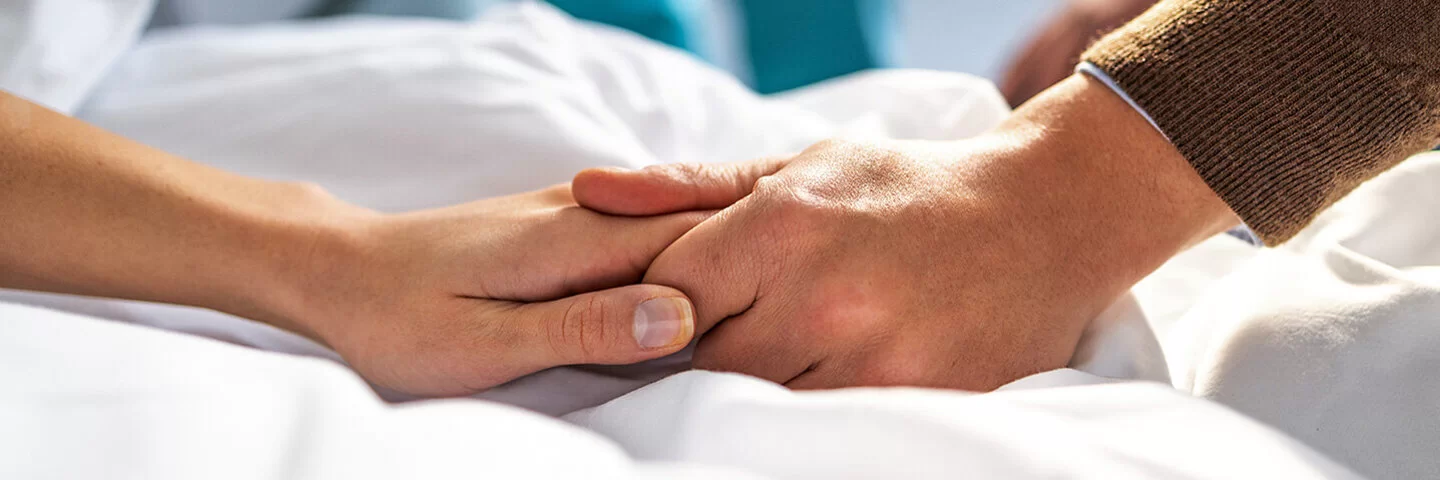 Eine Frau liegt in einem Krankenbett, ein Mann sitzt neben ihr. Zu sehen sind jedoch nur die Hände der beiden, die sich festhalten. Im Hintergrund eine medizinische Fachkraft in grüner Kleidung.