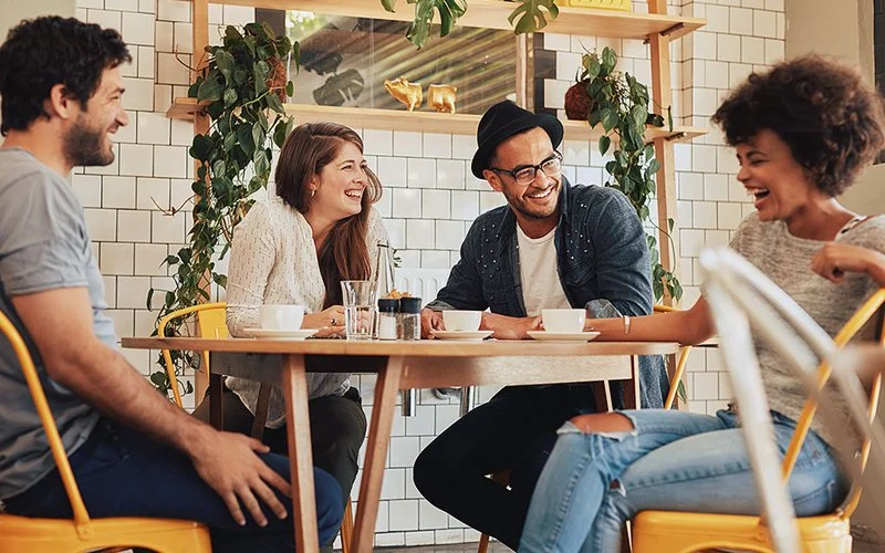 An einem Tisch in einem Café sitzt eine Gruppe junger Erwachsener: zwei Männer und zwei Frauen. Eine der Frauen lacht, die anderen drei blicken sie an und lächeln.