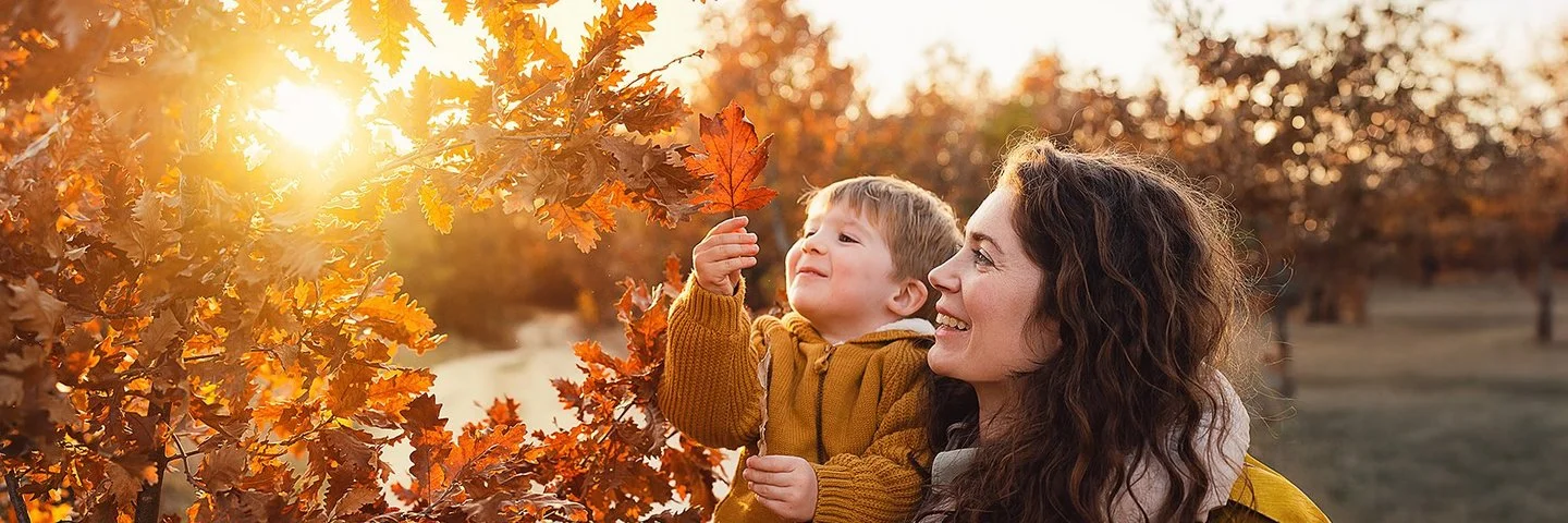 Eine junge Frau hält lächelnd einen etwa dreijährigen Jungen auf dem Arm, der ein rot verfärbtes Blatt von einer Eiche betrachtet. Durch den Baum scheint die untergehende Herbstsonne.