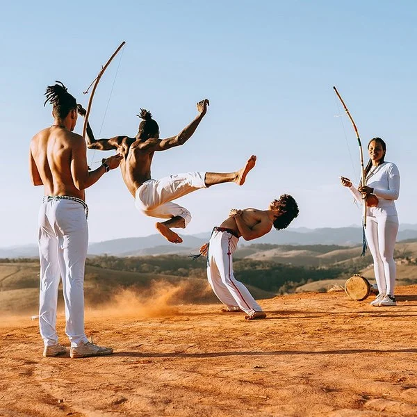 In einer wüstenähnlichen Landschaft springt ein junger Capoeirista hoch, ein anderer weicht ihm aus, indem er den Oberkörper nach hinten beut. Eine Frau und ein Mann feuern die beiden mit Musikinstrumenten an.