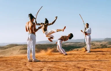 In einer wüstenähnlichen Landschaft springt ein junger Capoeirista hoch, ein anderer weicht ihm aus, indem er den Oberkörper nach hinten beut. Eine Frau und ein Mann feuern die beiden mit Musikinstrumenten an.