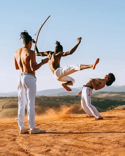 In einer wüstenähnlichen Landschaft springt ein junger Capoeirista hoch, ein anderer weicht ihm aus, indem er den Oberkörper nach hinten beut. Eine Frau und ein Mann feuern die beiden mit Musikinstrumenten an.