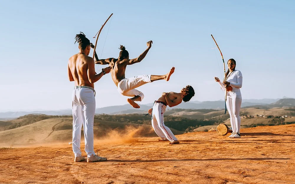 In einer wüstenähnlichen Landschaft springt ein junger Capoeirista hoch, ein anderer weicht ihm aus, indem er den Oberkörper nach hinten beut. Eine Frau und ein Mann feuern die beiden mit Musikinstrumenten an.