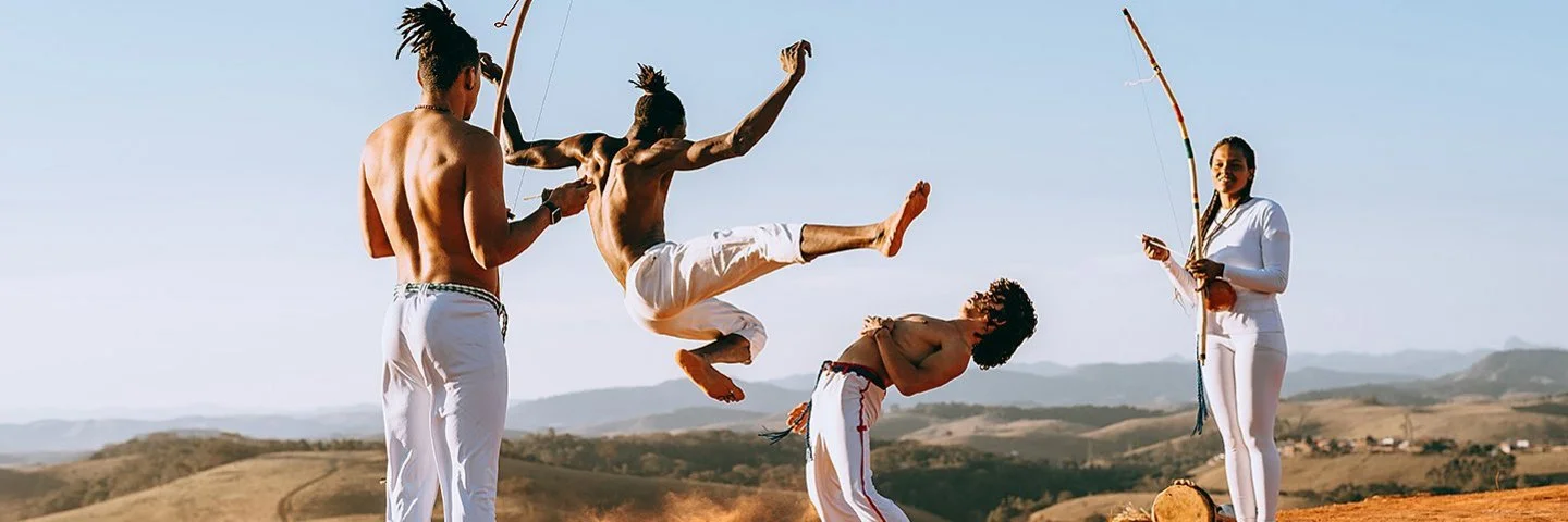 In einer wüstenähnlichen Landschaft springt ein junger Capoeirista hoch, ein anderer weicht ihm aus, indem er den Oberkörper nach hinten beut. Eine Frau und ein Mann feuern die beiden mit Musikinstrumenten an.