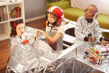 Drei Kinder im Grundschulalter spielen im Wohnzimmer mit einem selbstgebauten Raumschiff aus Pappe und Alufolie. Ein Mädchen mit Helm sitzt im Cockpit des Raumschiffs, hinter ihr ein Junge mit Headset und silberner Jacke. Ein jüngeres Mädchen mit großer Schutzbrille und ebenfalls silberner Jacke kniet neben dem Raumschiff.