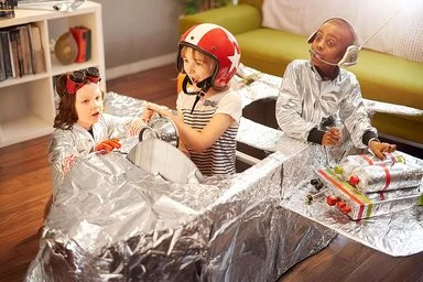 Drei Kinder im Grundschulalter spielen im Wohnzimmer mit einem selbstgebauten Raumschiff aus Pappe und Alufolie. Ein Mädchen mit Helm sitzt im Cockpit des Raumschiffs, hinter ihr ein Junge mit Headset und silberner Jacke. Ein jüngeres Mädchen mit großer Schutzbrille und ebenfalls silberner Jacke kniet neben dem Raumschiff.