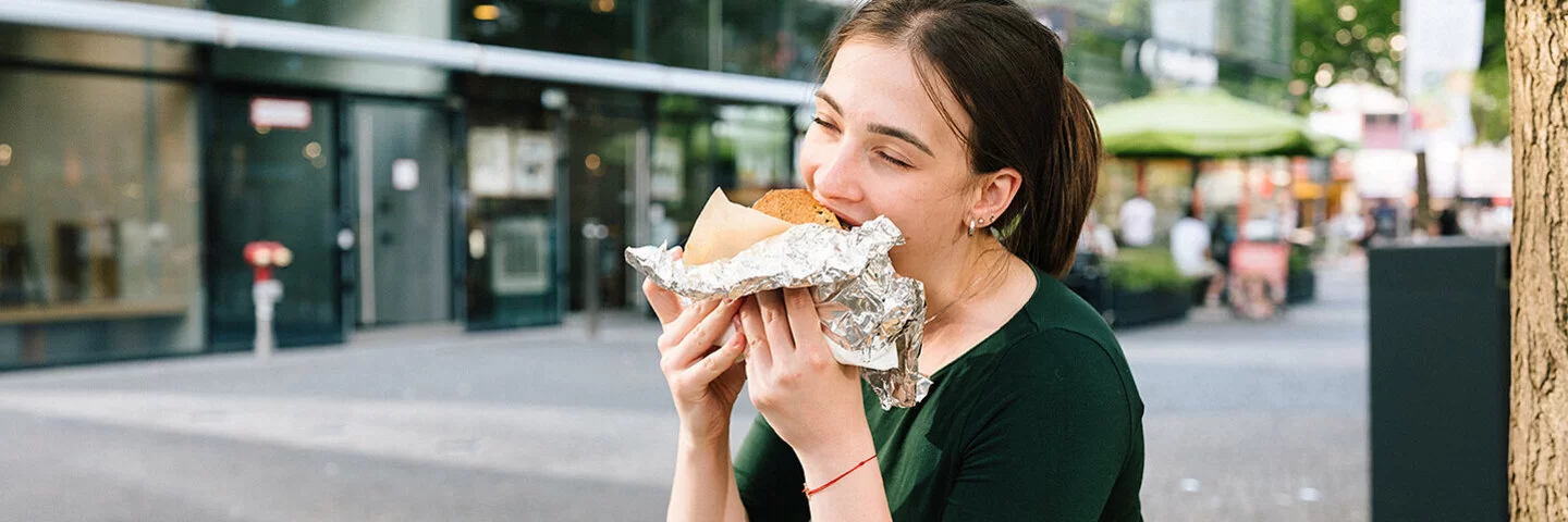 Eine junge Frau sitzt in einer Einkaufsstraße und isst ein großes gefülltes Stück Brot, das halb in Alufolie eingepackt ist.