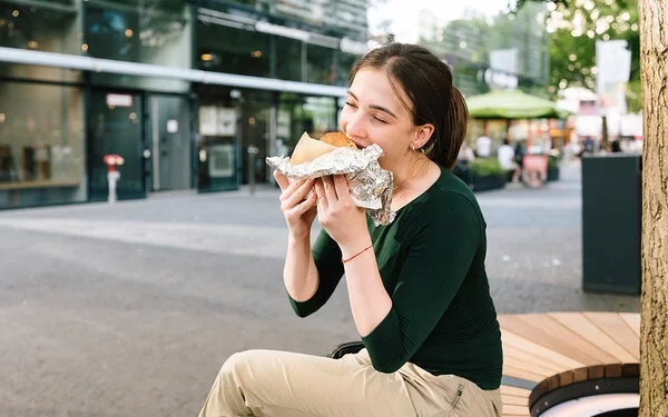 Eine junge Frau sitzt in einer Einkaufsstraße und isst ein großes gefülltes Stück Brot, das halb in Alufolie eingepackt ist.