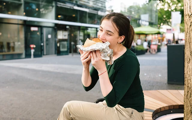 Eine junge Frau sitzt in einer Einkaufsstraße und isst ein großes gefülltes Stück Brot, das halb in Alufolie eingepackt ist.