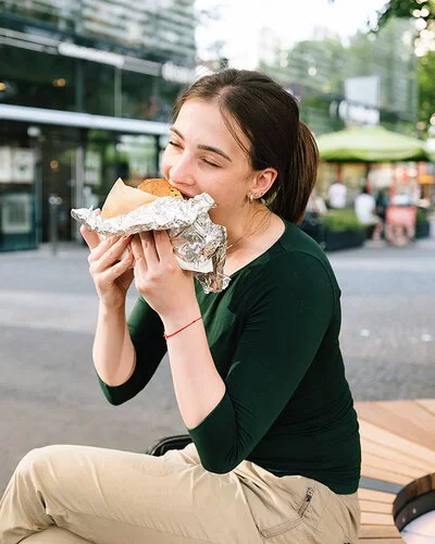 Eine junge Frau sitzt in einer Einkaufsstraße und isst ein großes gefülltes Stück Brot, das halb in Alufolie eingepackt ist.