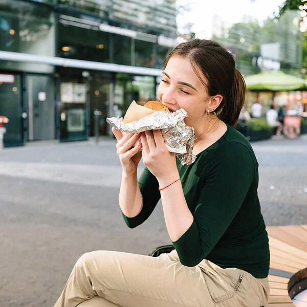 Eine junge Frau sitzt in einer Einkaufsstraße und isst ein großes gefülltes Stück Brot, das halb in Alufolie eingepackt ist.