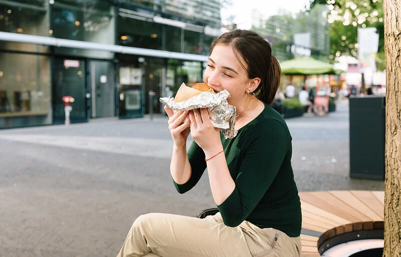Eine junge Frau sitzt in einer Einkaufsstraße und isst ein großes gefülltes Stück Brot, das halb in Alufolie eingepackt ist.
