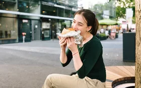 Eine junge Frau sitzt in einer Einkaufsstraße und isst ein großes gefülltes Stück Brot, das halb in Alufolie eingepackt ist.