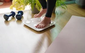 Eine Person steigt auf eine Körperfettwaage. Rechts und links von der Waage liegen eine ausgebreitete Sportmatte und Hanteln.
