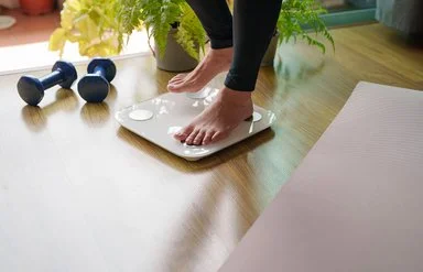 Eine Person steigt auf eine Körperfettwaage. Rechts und links von der Waage liegen eine ausgebreitete Sportmatte und Hanteln.
