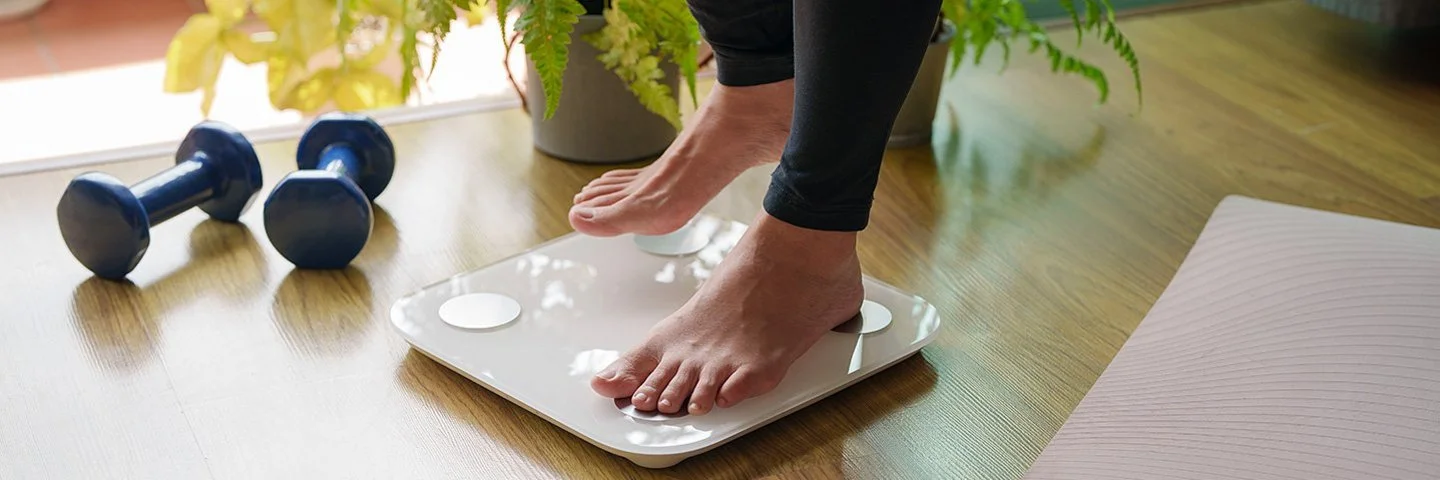 Eine Person steigt auf eine Körperfettwaage. Rechts und links von der Waage liegen eine ausgebreitete Sportmatte und Hanteln.
