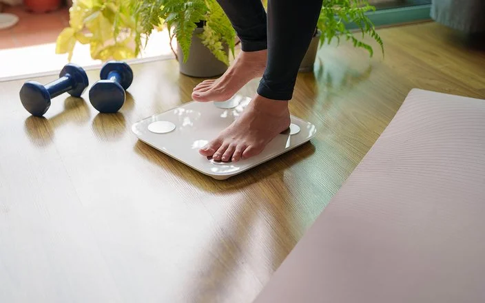 Eine Person steigt auf eine Körperfettwaage. Rechts und links von der Waage liegen eine ausgebreitete Sportmatte und Hanteln.