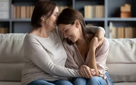 Eine Mutter umarmt ihre jugendliche Tochter auf einem Sofa verständnisvoll. Beide lächeln.