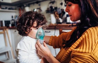 Ein kleiner Junge inhaliert zuhause. Seine Mutter hält ihm die Inhalationsmaske über Mund und Nase.