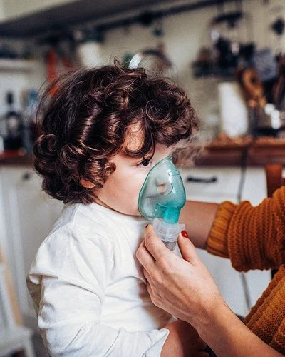 Ein kleiner Junge inhaliert zuhause. Seine Mutter hält ihm die Inhalationsmaske über Mund und Nase.