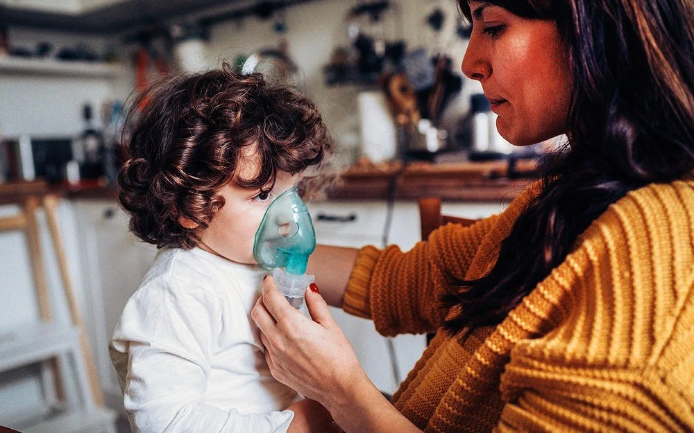 Ein kleiner Junge inhaliert zuhause. Seine Mutter hält ihm die Inhalationsmaske über Mund und Nase.