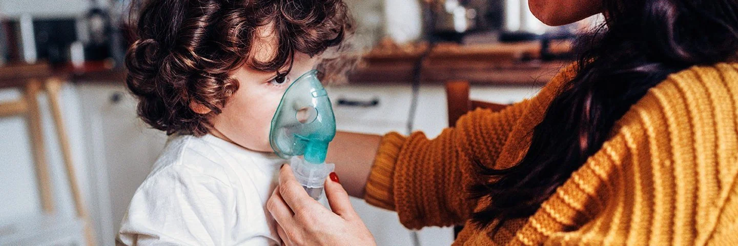 Ein kleiner Junge inhaliert zuhause. Seine Mutter hält ihm die Inhalationsmaske über Mund und Nase.