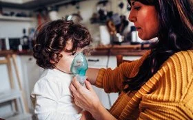 Ein kleiner Junge inhaliert zuhause. Seine Mutter hält ihm die Inhalationsmaske über Mund und Nase.