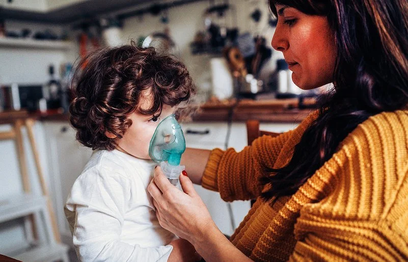 Ein kleiner Junge inhaliert zuhause. Seine Mutter hält ihm die Inhalationsmaske über Mund und Nase.