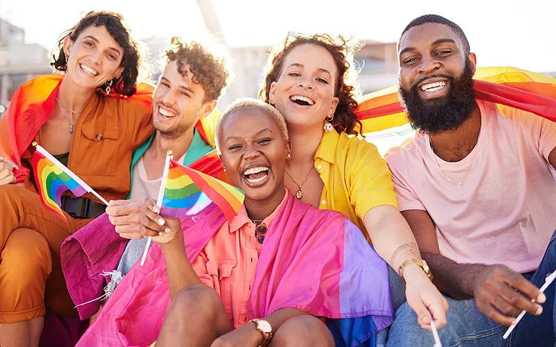Eine Gruppe von Menschen unterschiedlichen Geschlechts und Herkunft in bunter Kleidung mit der LGBTQ-Regenbogen-Fahne.