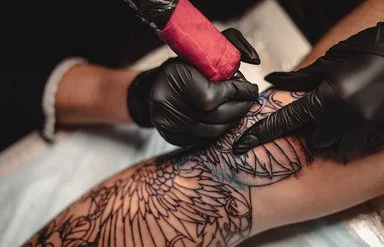 Eine nicht sichtbare Person mit schwarzen Schutzhandschuhen sticht mit einer Tätowiermaschine ein großes und aufwändiges Tattoo auf einem Bein.